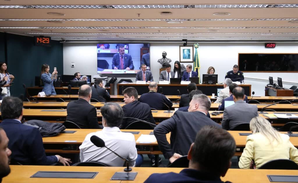 Audiência pública em plenário com participantes, painel de monitoramento e apresentação de fatos.